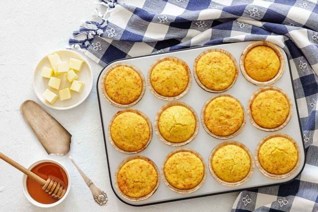 A muffin tin with twelve baked cornbread muffins sits on a table next to a bowl of butter cubes, a dish of honey with a dipper, and a blue checkered cloth.