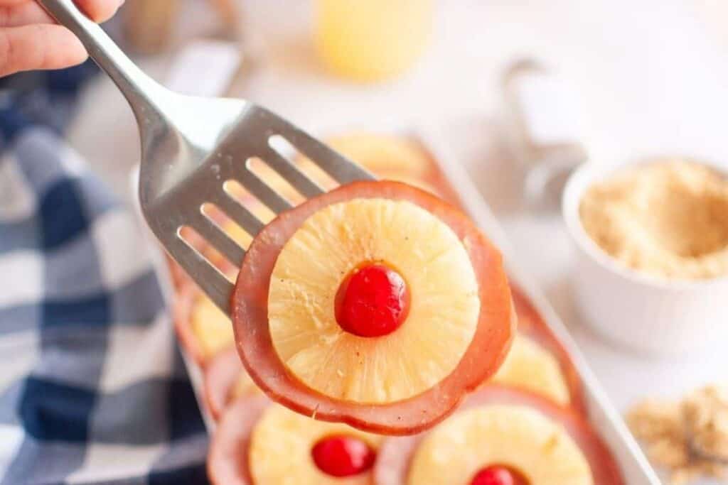 A metal spatula holds a slice of ham topped with a pineapple ring and a maraschino cherry. A tray with more slices and a bowl of brown sugar are in the background.