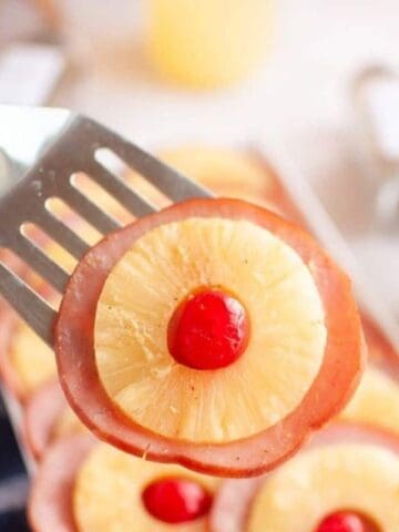 A metal spatula holds a slice of ham topped with a pineapple ring and a maraschino cherry. A tray with more slices and a bowl of brown sugar are in the background.