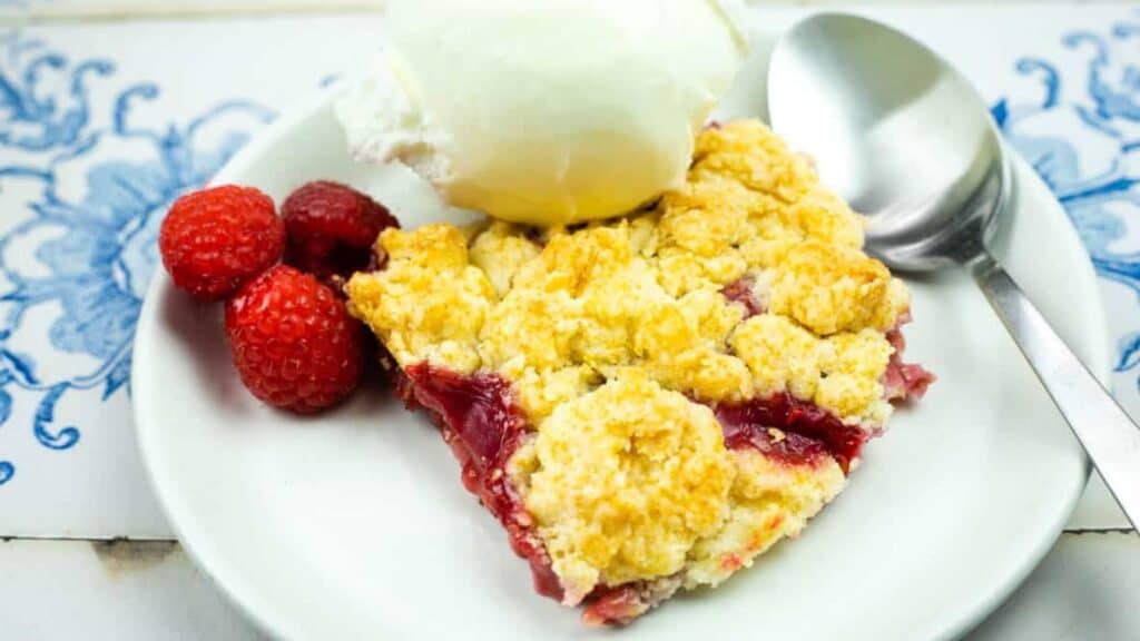 A plate with a slice of fruit crumble topped with a scoop of vanilla ice cream, three raspberries on the side, and a spoon on the plate.