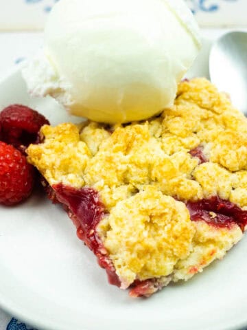 A plate with a slice of fruit crumble, a scoop of vanilla ice cream, three raspberries, and a spoon on a white plate.