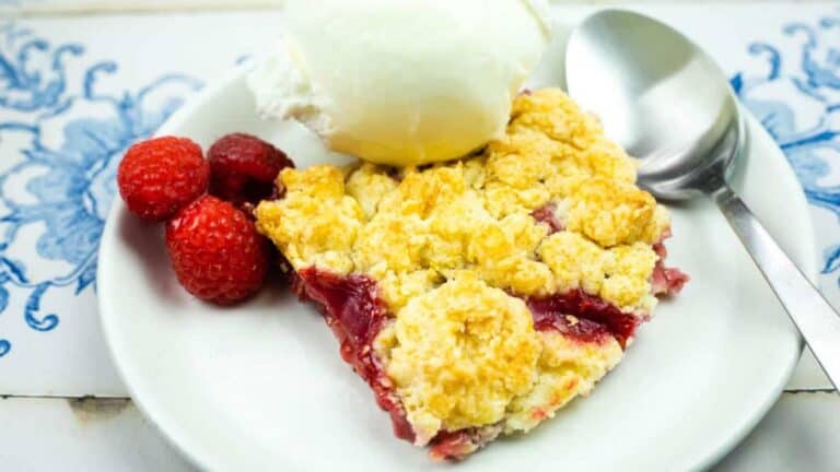 A plate with a slice of fruit crumble topped with a scoop of vanilla ice cream, three raspberries on the side, and a spoon on the plate.