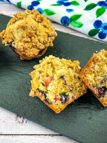 Four blueberry muffins, one cut in half to show the inside, are displayed on a black slate platter with a blue and green floral cloth in the background.