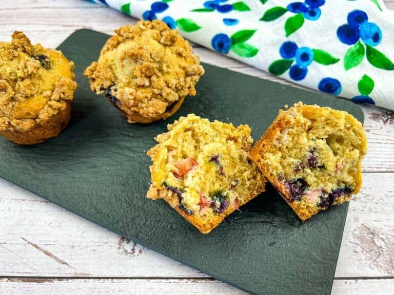 Four blueberry muffins, one cut in half to show the inside, are displayed on a black slate platter with a blue and green floral cloth in the background.