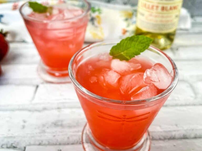 Two glasses of a pink iced beverage garnished with mint leaves are placed on a light wooden surface, with a bottle in the background.