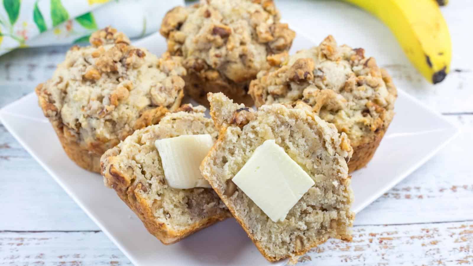 Banana nut muffins on a white plate, one muffin split open with two pats of butter on top, and a banana in the background.