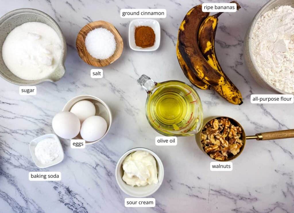 Ingredients for banana bread arranged on a marble surface, including sugar, salt, ground cinnamon, ripe bananas, flour, eggs, baking soda, olive oil, walnuts, and sour cream.
