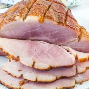 Sliced glazed ham arranged on a white plate, with a floral napkin and an orange flower in the background.