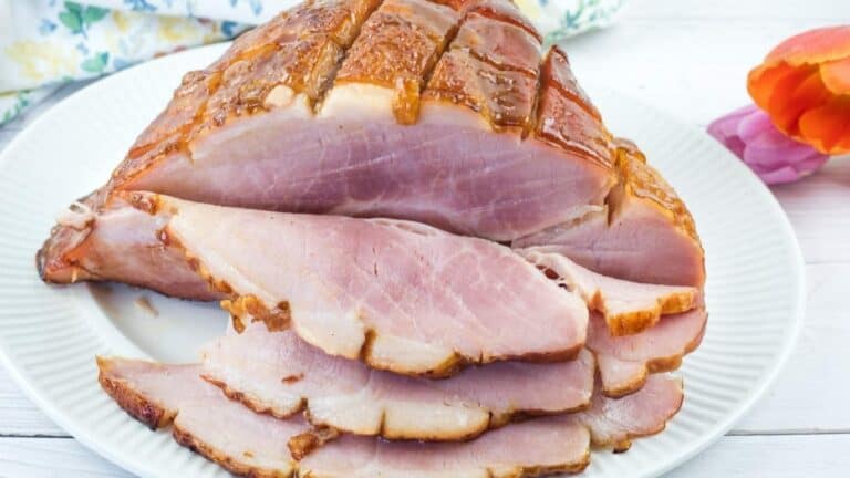 Sliced glazed ham arranged on a white plate, with a floral napkin and an orange flower in the background.