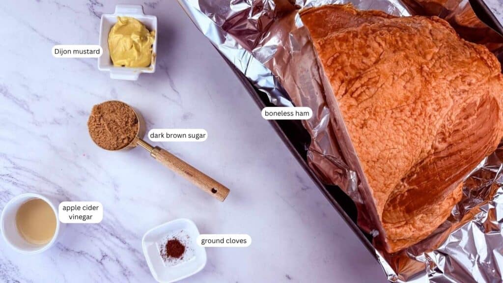 A boneless ham in a foil-lined pan with labeled ingredients: Dijon mustard, dark brown sugar, apple cider vinegar, and ground cloves on a marble surface.