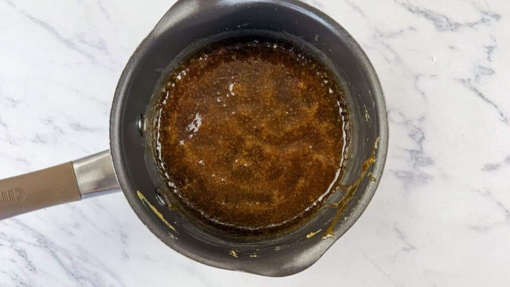 Overhead view of a saucepan containing a brown, bubbling liquid mixture on a white marble surface.