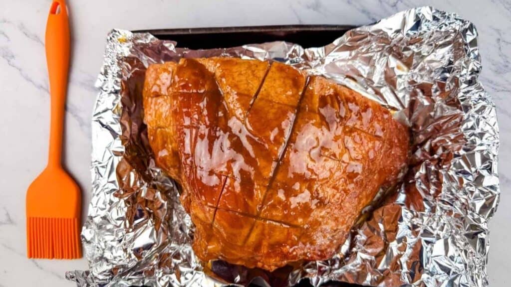 A glazed ham with crosshatched cuts rests on aluminum foil next to an orange silicone basting brush on a marble surface.