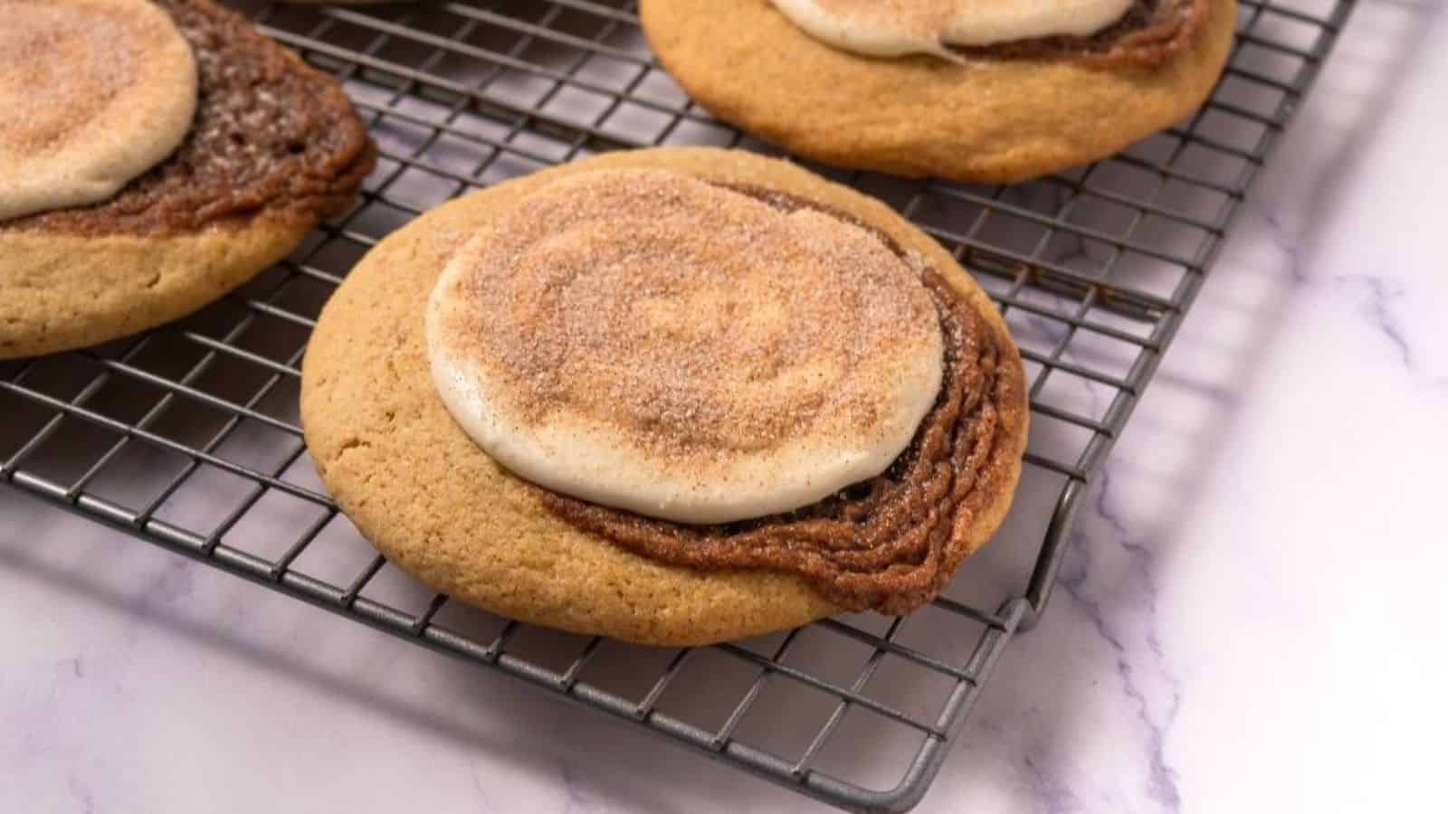 Close-up of cinnamon roll cookies with icing and cinnamon sugar on top, cooling on a wire rack over a marble surface.