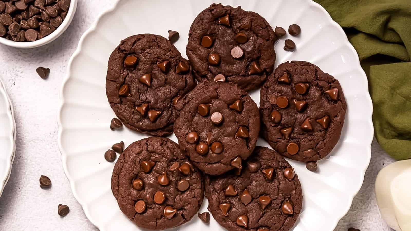 Six Copycat Crumbl Dark Dream Cookies are arranged on a white scalloped plate, with extra chocolate chips scattered around.