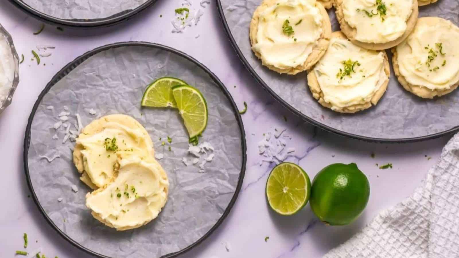 Plates of frosted cookies garnished with lime zest, with lime slices and a whole lime on a marble surface.
