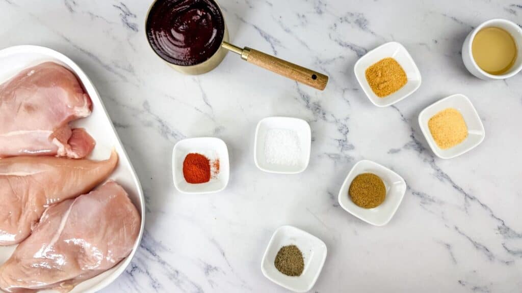 Raw chicken breasts on a plate, small bowls with various spices, a bowl of liquid marinade, and a pot of sauce arranged on a marble surface.