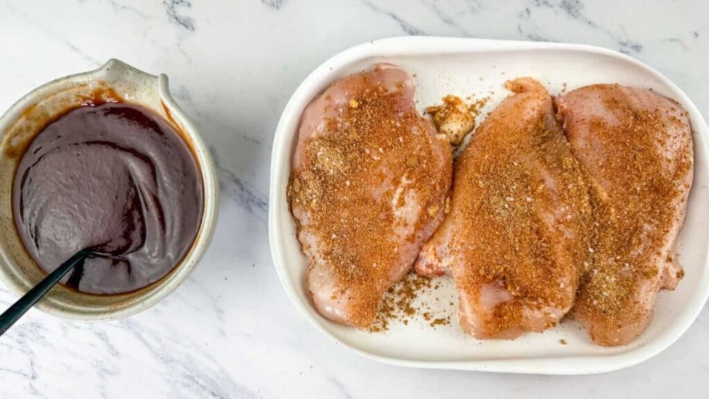Three raw chicken breasts coated with dry seasoning on a white plate next to a bowl of thick, dark barbecue sauce with a spoon, all on a white marble surface.