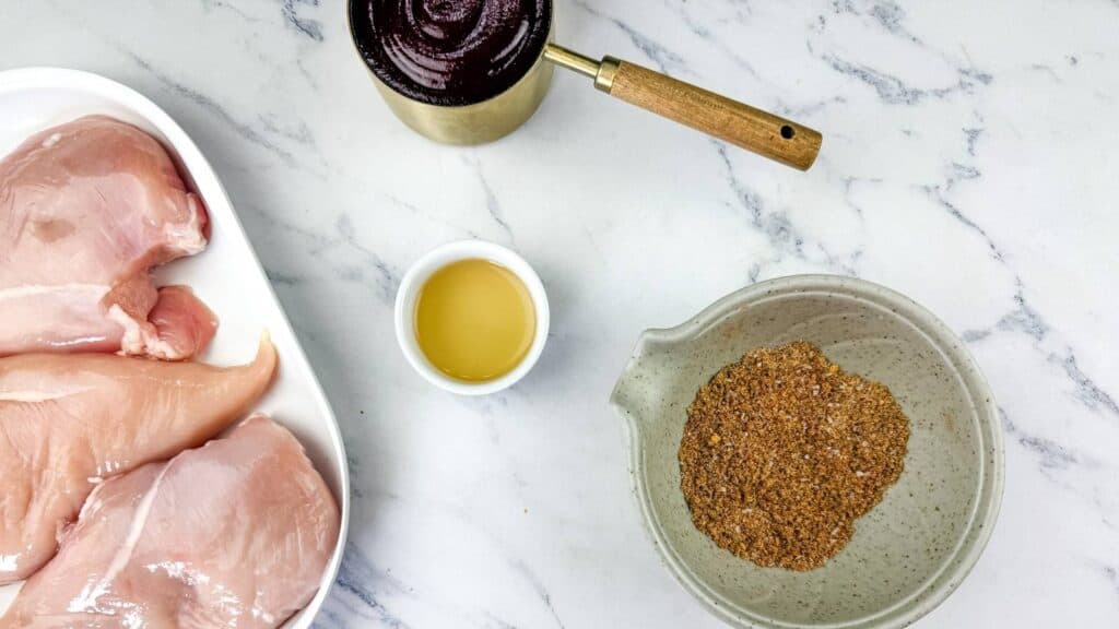 Raw chicken breasts on a white tray, a small bowl of oil, a pot of dark liquid, and a bowl of dry seasoning on a marble countertop.