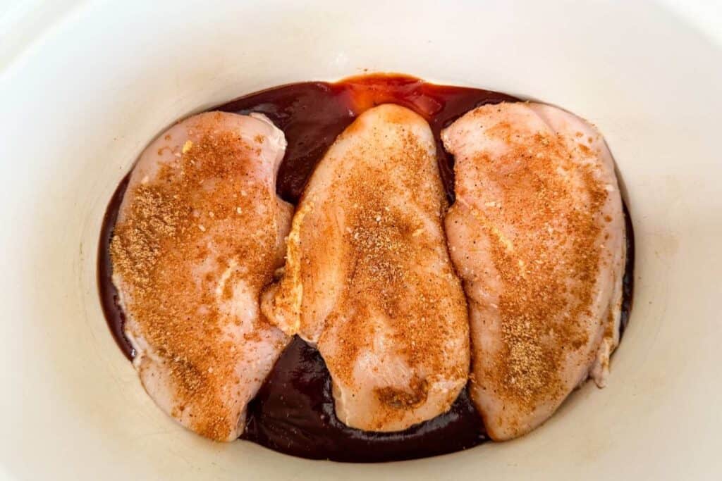 Three raw chicken breasts seasoned with spices are placed on top of a layer of barbecue sauce in a white dish.