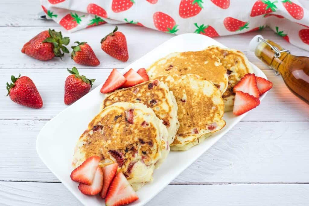 Three strawberry pancakes with sliced strawberries are arranged on a white plate, surrounded by whole strawberries and a glass syrup bottle on a white wooden table.