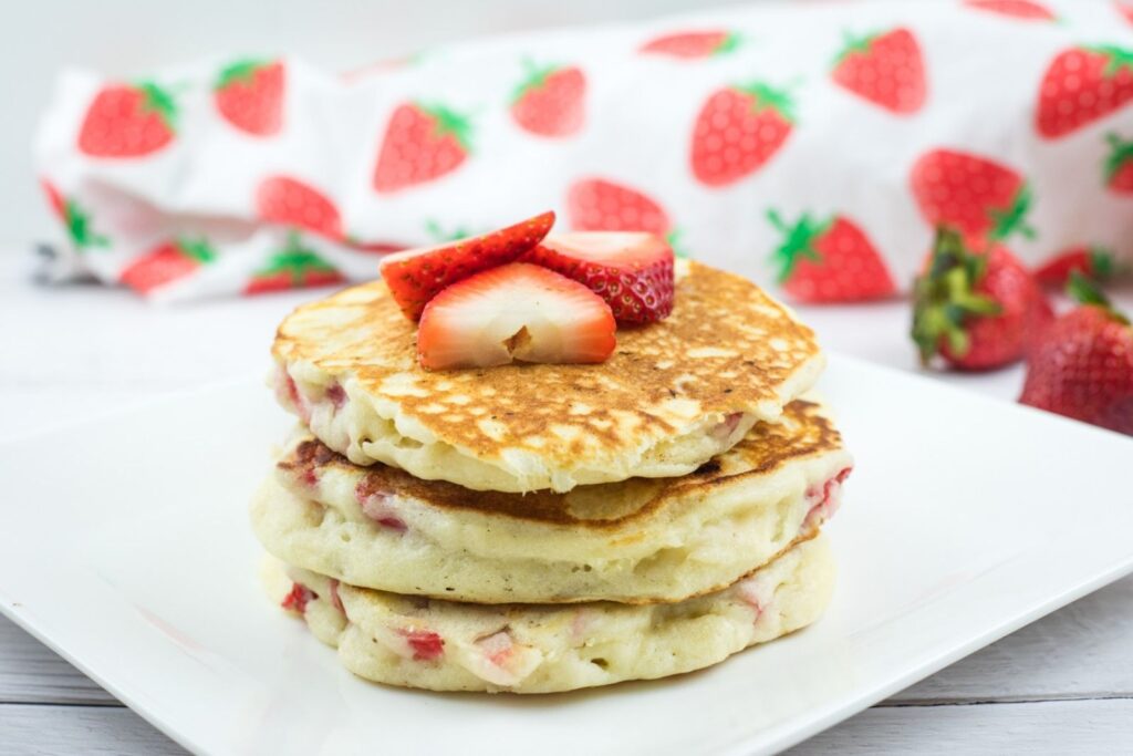 A stack of three pancakes with strawberry pieces, topped with sliced strawberries, on a white plate; a strawberry-patterned cloth is in the background.