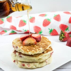 A hand pours syrup onto a stack of pancakes topped with sliced strawberries, with whole strawberries and a strawberry-patterned cloth in the background.