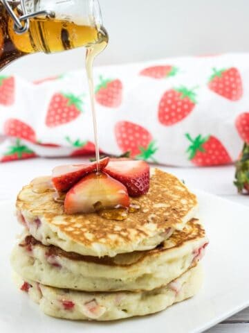 A hand pours syrup onto a stack of pancakes topped with sliced strawberries, with whole strawberries and a strawberry-patterned cloth in the background.