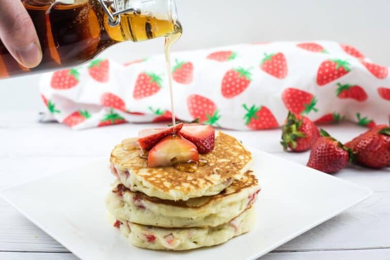 A hand pours syrup onto a stack of pancakes topped with sliced strawberries, with whole strawberries and a strawberry-patterned cloth in the background.