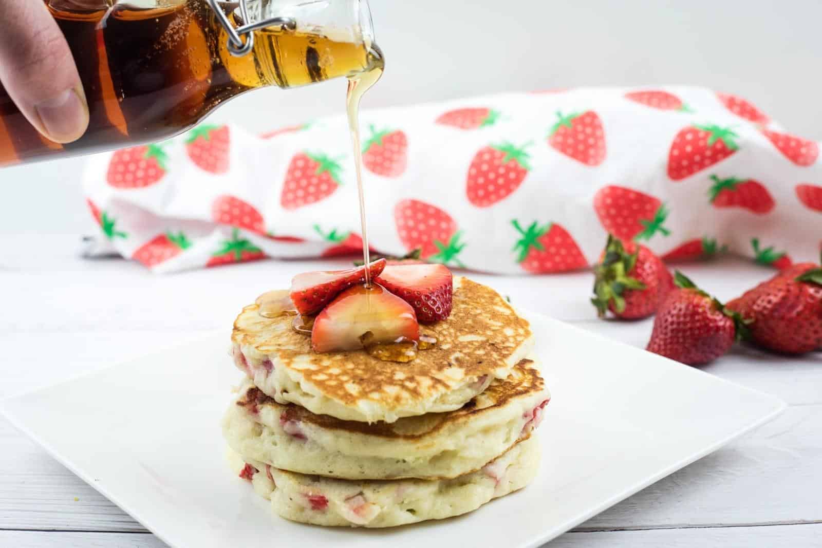 A hand pours syrup onto a stack of pancakes topped with sliced strawberries, with whole strawberries and a strawberry-patterned cloth in the background.