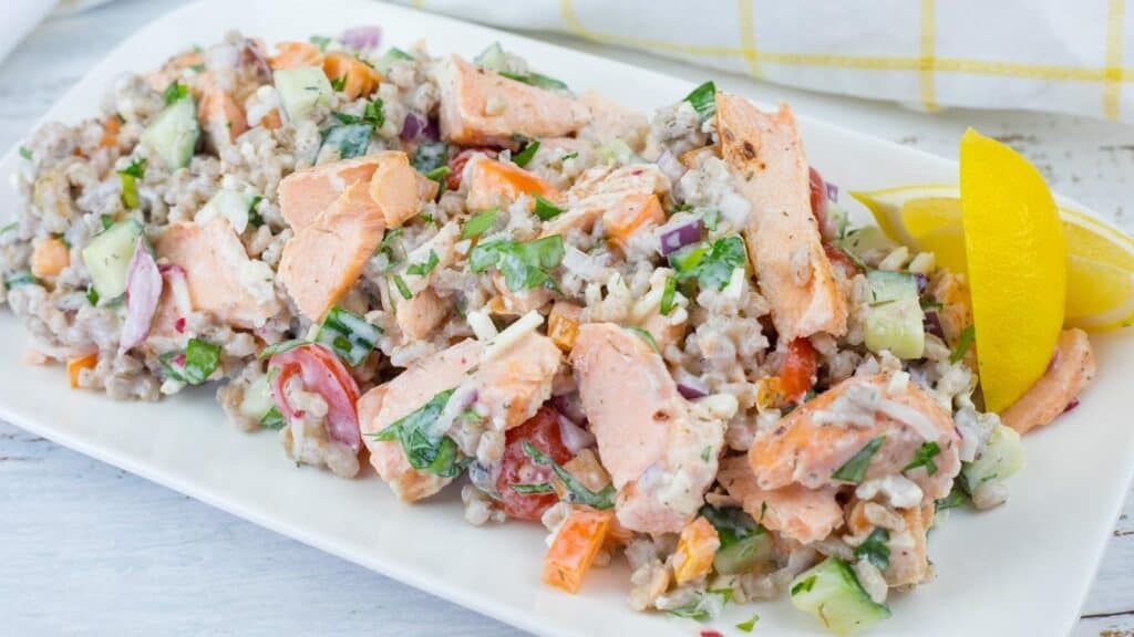 A rectangular white plate of Mediterranean Salmon Salad with Barley, topped with fresh herbs, and a lemon wedge on the side.