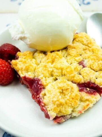 A dessert plate with a slice of berry crumble, a scoop of vanilla ice cream, three raspberries, and a spoon on a white and blue patterned table.