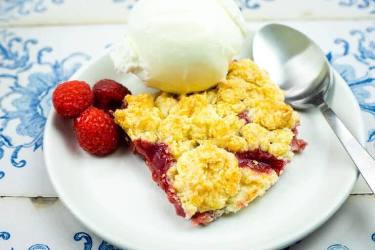 A dessert plate with a slice of berry crumble, a scoop of vanilla ice cream, three raspberries, and a spoon on a white and blue patterned table.