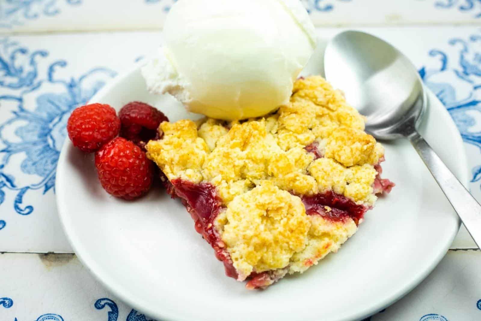 A dessert plate with a slice of berry crumble, a scoop of vanilla ice cream, three raspberries, and a spoon on a white and blue patterned table.
