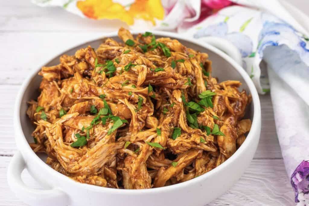 A white bowl filled with shredded barbecue chicken garnished with chopped parsley, placed on a light wooden surface next to a colorful napkin.