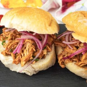 Two pulled chicken sandwiches topped with sliced red onions on crusty buns, served on a black slate plate with a side of grape tomatoes.