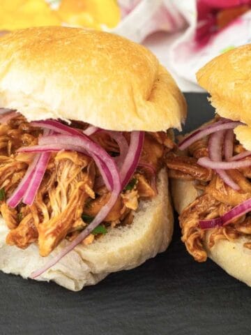 Two pulled chicken sandwiches topped with sliced red onions on crusty buns, served on a black slate plate with a side of grape tomatoes.