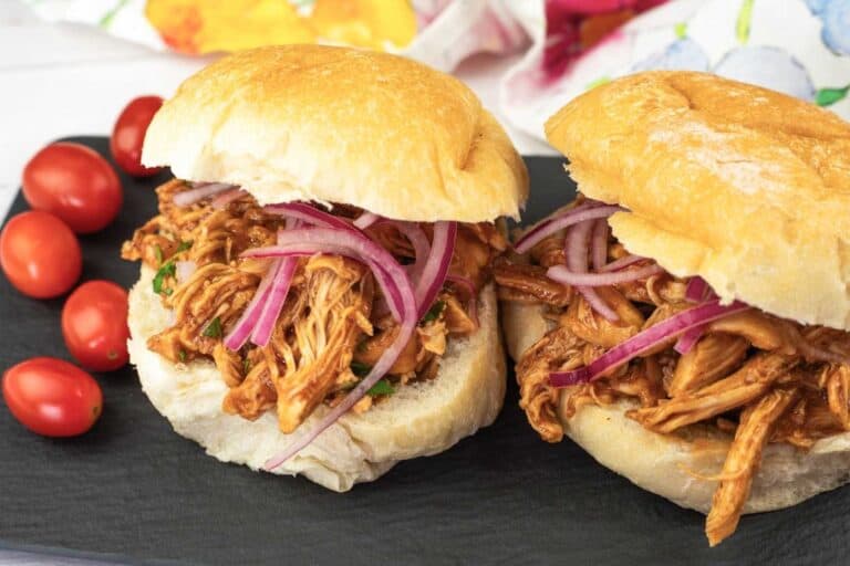 Two pulled chicken sandwiches topped with sliced red onions on crusty buns, served on a black slate plate with a side of grape tomatoes.