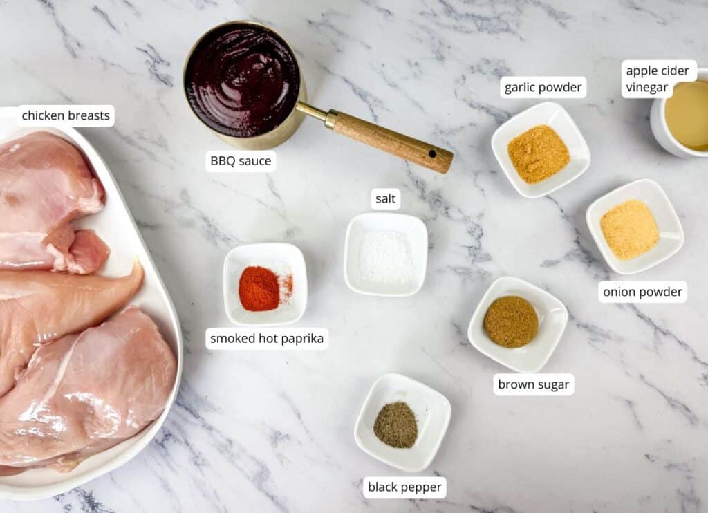 Raw chicken breasts, BBQ sauce, small bowls of spices (smoked paprika, salt, black pepper, onion powder, garlic powder), brown sugar, and apple cider vinegar on a white surface.