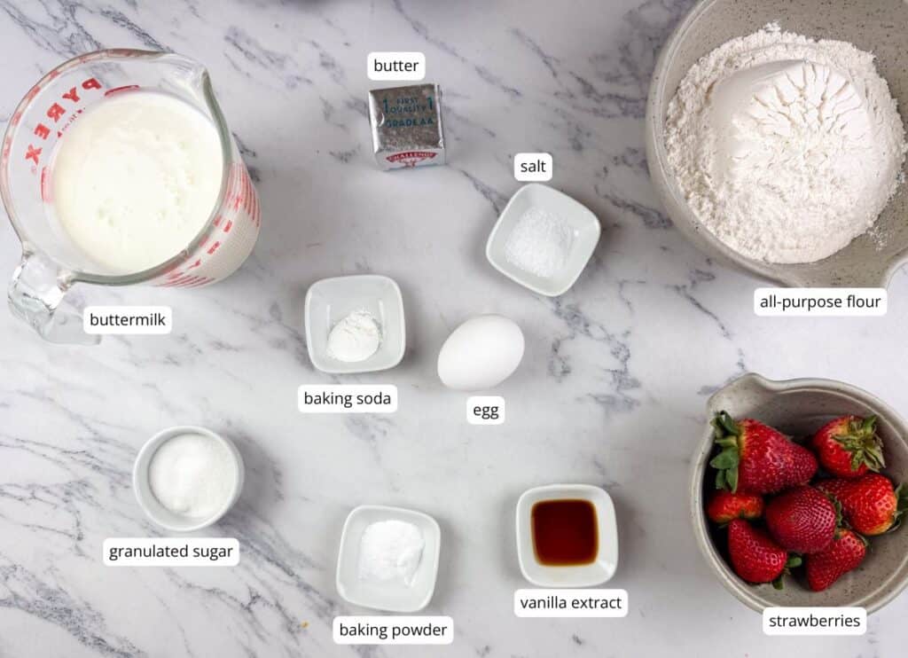 An overhead view of labeled baking ingredients on a marble surface, including buttermilk, butter, salt, flour, baking soda, egg, sugar, baking powder, vanilla extract, and strawberries.
