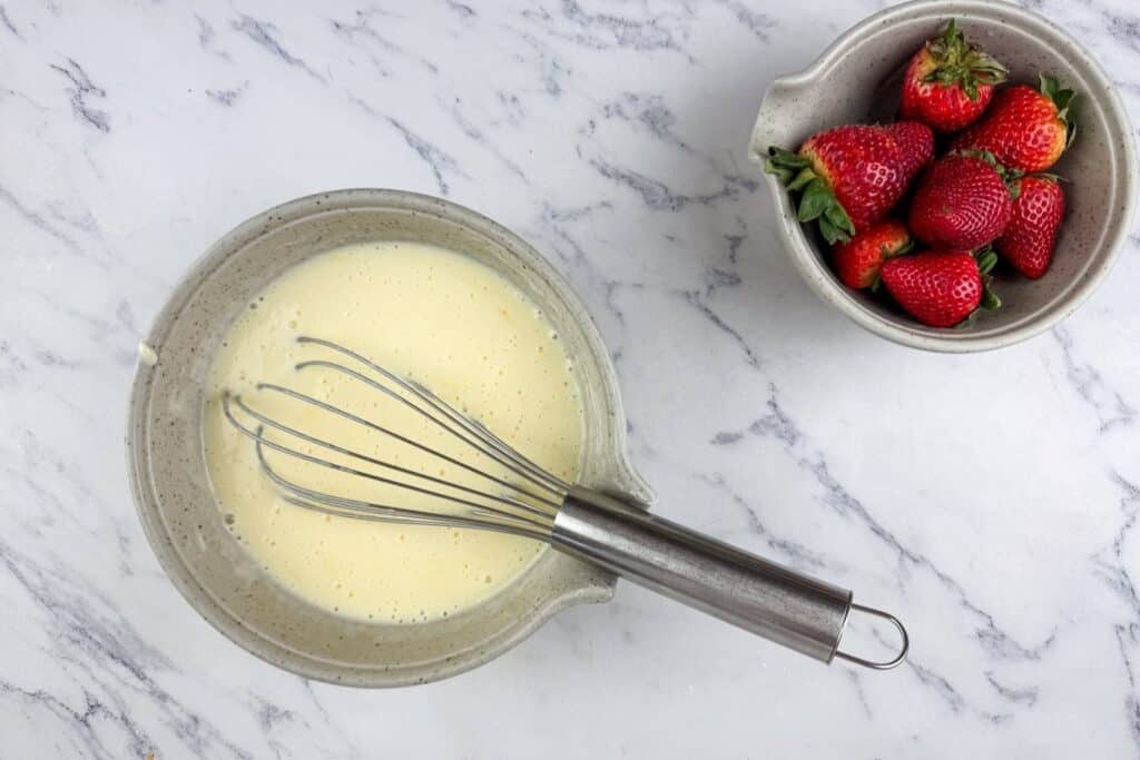 A whisk rests in a bowl of batter next to a smaller bowl filled with fresh strawberries on a marble surface.