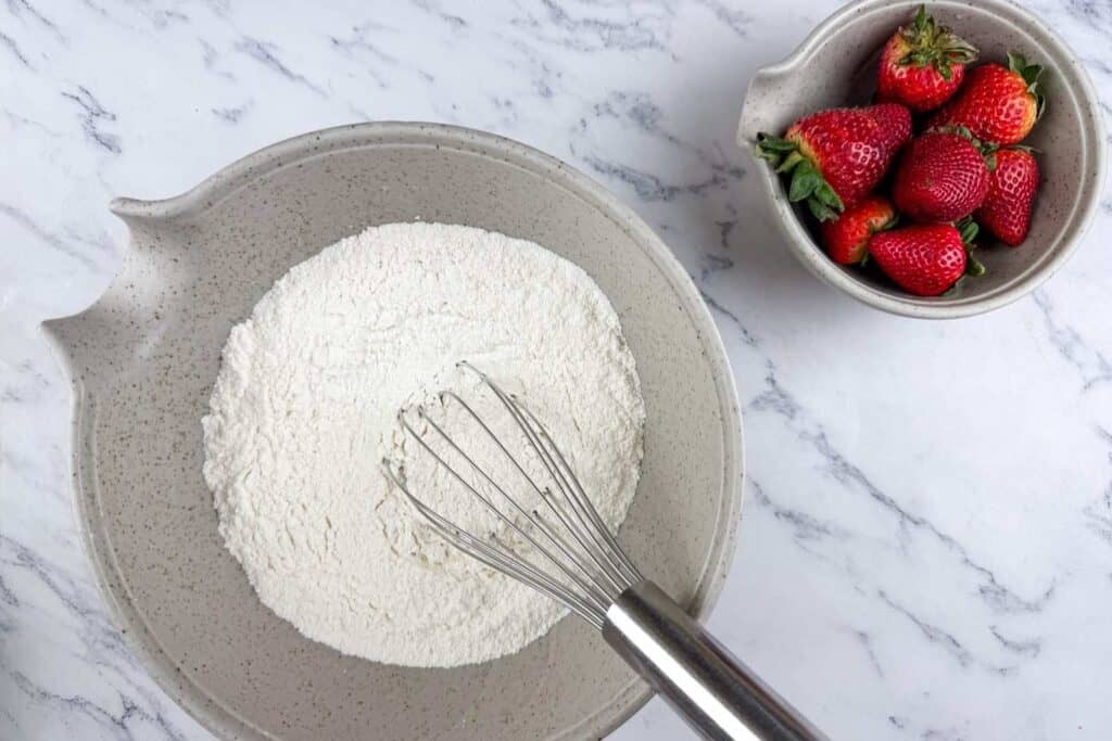A bowl of flour with a whisk inside sits next to a bowl of fresh strawberries on a white marble surface.