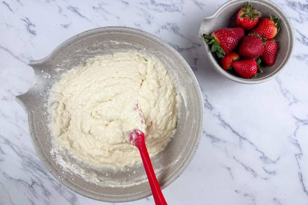 A mixing bowl with batter and a red spatula next to a bowl of fresh strawberries on a marble countertop.