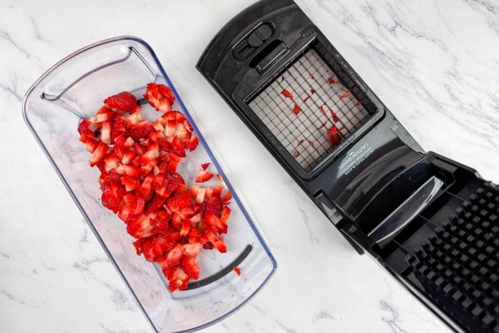 Chopped strawberries in a clear container next to a black vegetable chopper on a white marble surface.