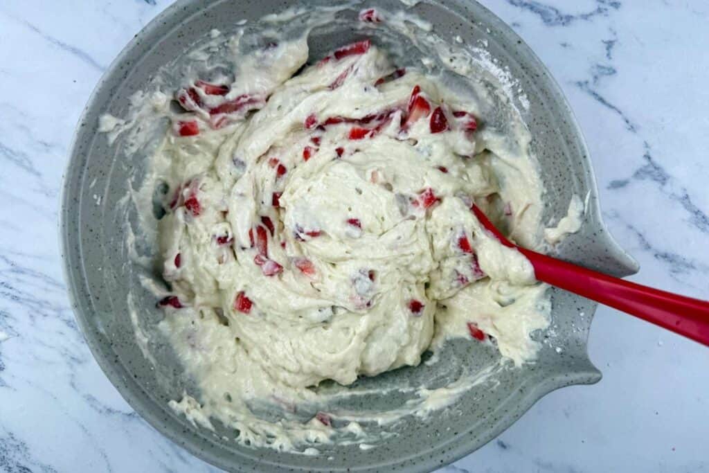 A mixing bowl containing thick batter with visible red fruit pieces and a red spatula on a marble surface.