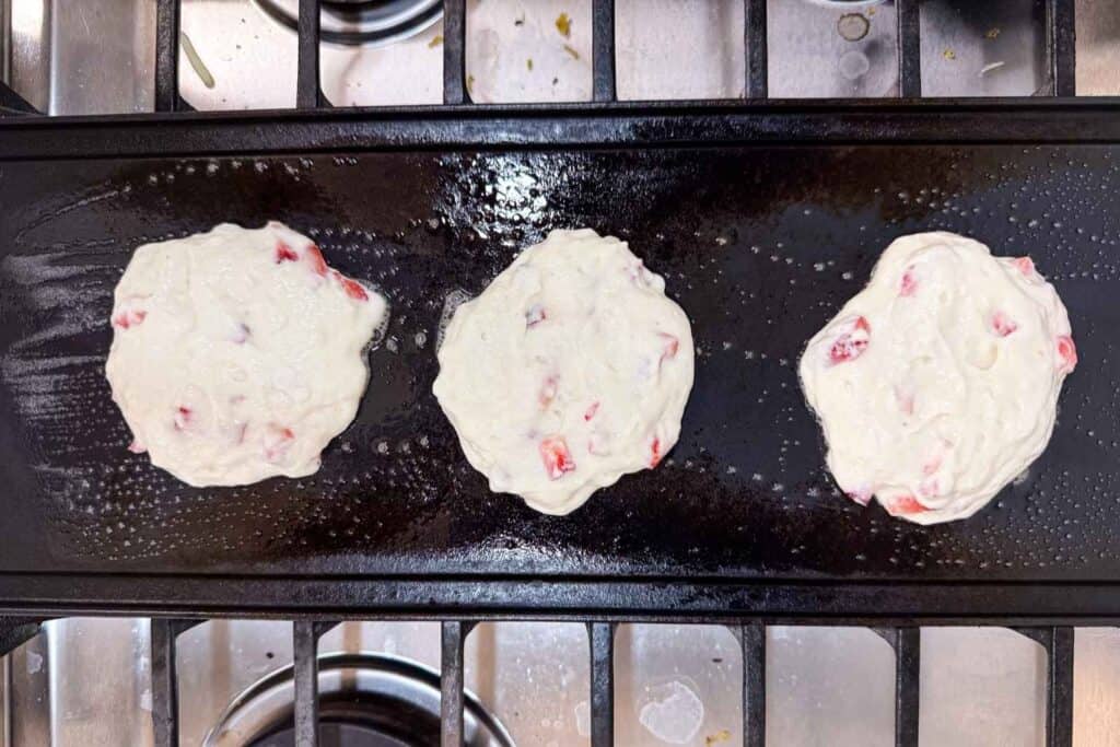 Three uncooked pancakes with visible red pieces on a black griddle over a stovetop.