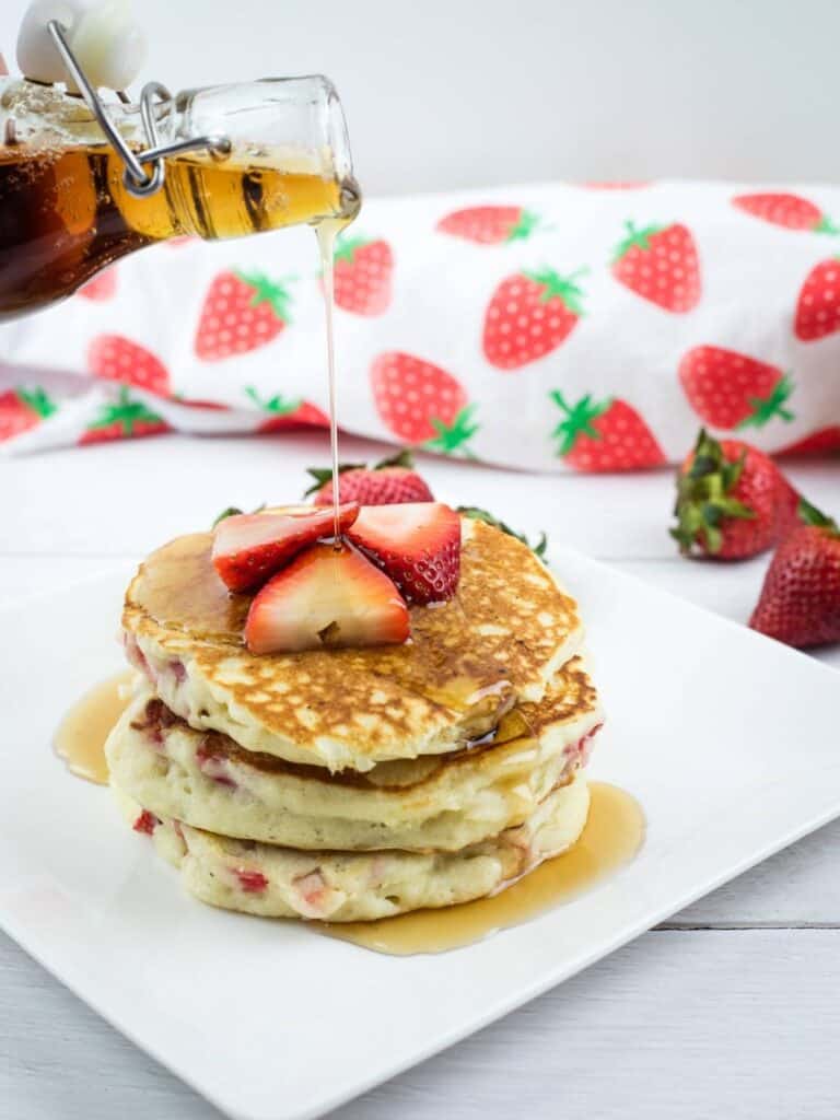 A stack of pancakes with sliced strawberries on top is being drizzled with syrup, with whole strawberries and a strawberry-patterned cloth in the background.