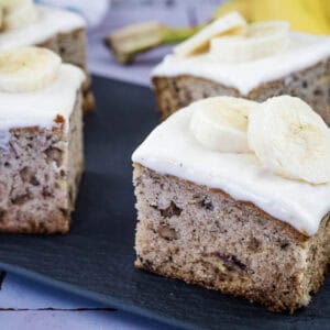 Four square pieces of banana cake with white frosting and banana slices on top are displayed on a dark serving tray.