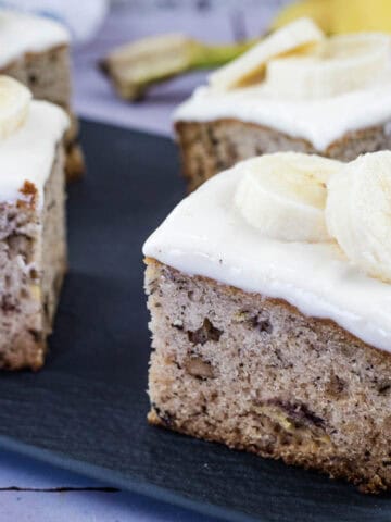 Four square pieces of banana cake with white frosting and banana slices on top are displayed on a dark serving tray.