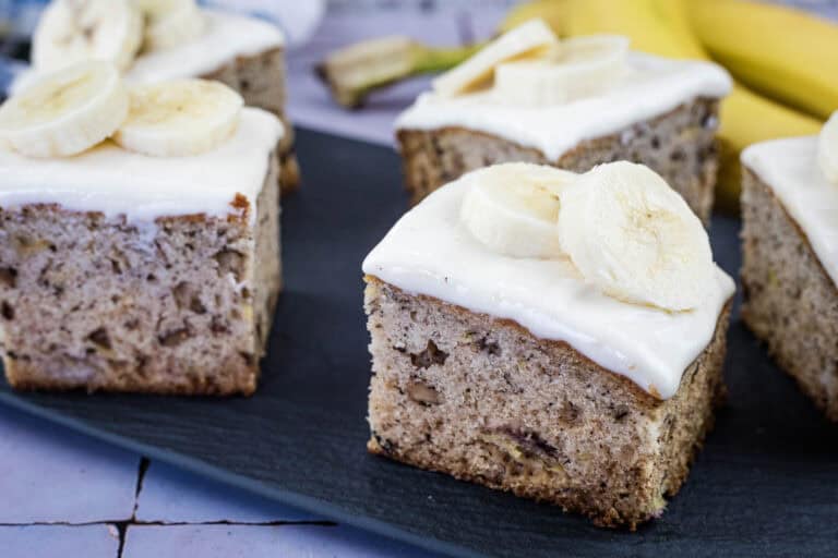 Four square pieces of banana cake with white frosting and banana slices on top are displayed on a dark serving tray.