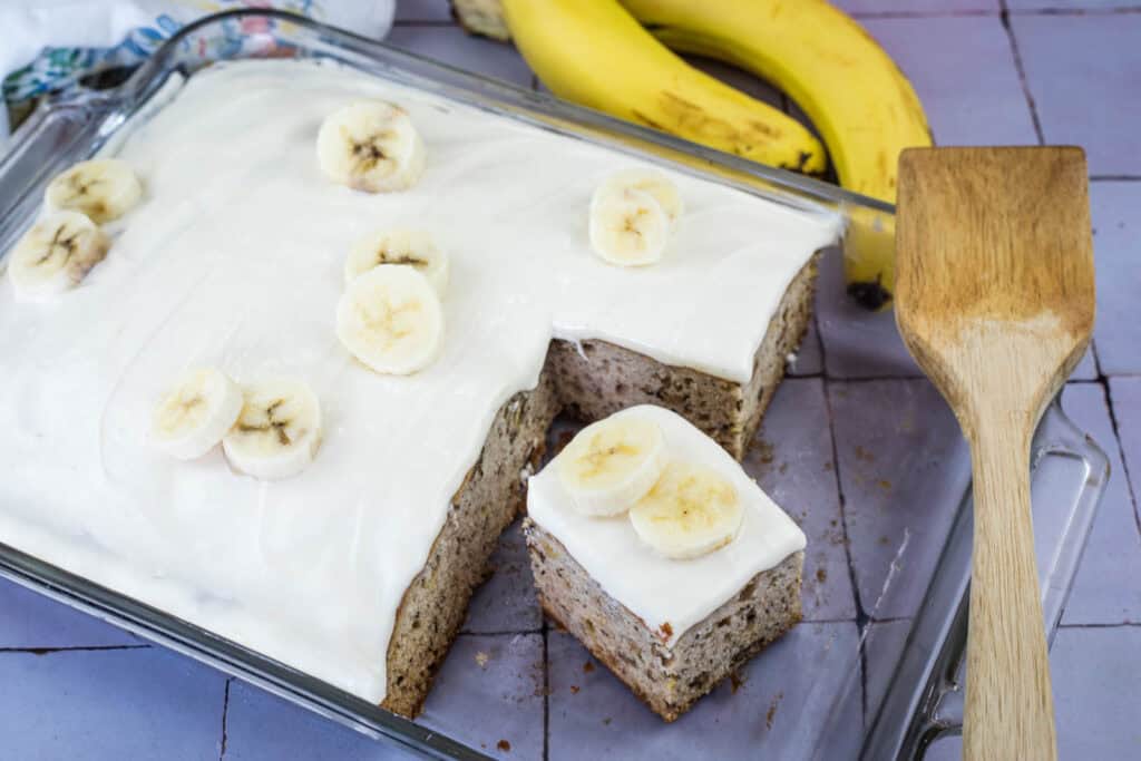 Glass baking dish with frosted banana cake, topped with banana slices. A slice is cut out and placed near a wooden spatula; two whole bananas are in the background.